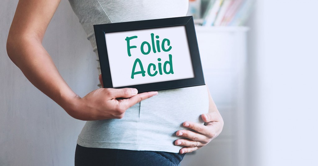 Pregnant woman holds whiteboard with text message - FOLIC ACID. Pregnancy, parenthood, preparation and expectation concept. Close-up, copy space, indoors; blog: Folic Acid During Pregnancy
