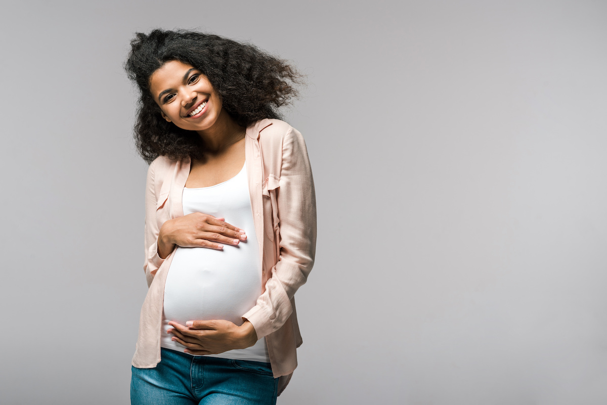 happy young pregnant african american woman touching belly on grey / blog - myths about pregnancy