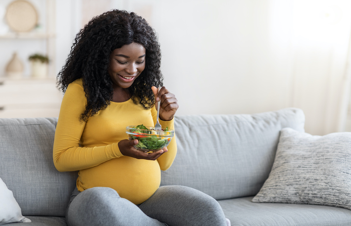 Hungry pregnant african american woman eating fresh salad for lunch, sitting on sofa in living room and looking at herHappy black woman expecting baby, enjoying healthy food; blog: 8 Tips for Great Pregnancy Nutrition