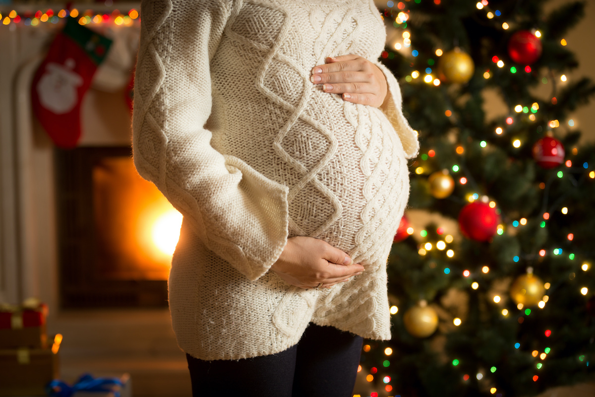 Closeup photo of pregnant woman posing against fireplace and Christmas tree; blog: Pregnancy Tips to Get You Through the Holiday Season