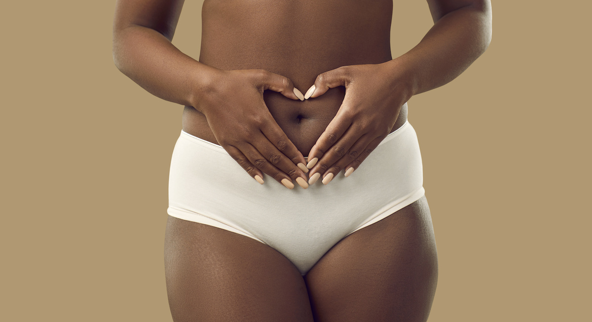 Black woman in underwear holding hands on abdominal area forming heart shape around navel, isolated on solid brown color background, cropped shot. Self love, care, health, pregnancy concept; blog: Yeast Infections